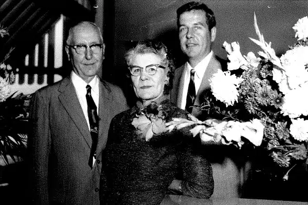 Allan Magnus "A.M." Anderson, Carrie Anderson & Richard "Dick" Anderson standing together next to a bouquet of flowers (1966 Grand Marais State Bank's 50th Anniversary)