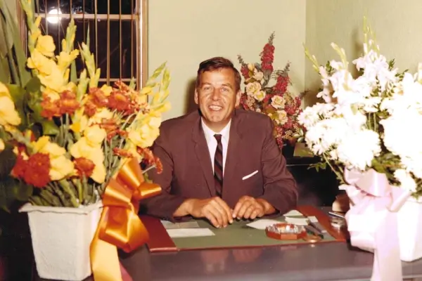 Richard "Dick" Anderson smiling sitting at a desk surrounded by flowers (1966 Grand Marais State Bank's 50th Anniversary)