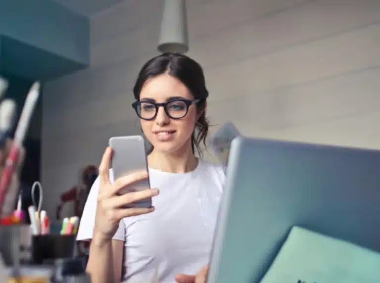 a young woman with glasses sitting in front of a laptop smiling at a smartphone with office supplies around her and an office in the background