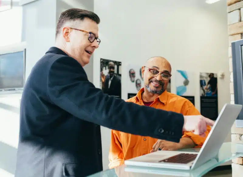 A man pointing at a laptop screen while another man watches and smiles next to him in a business setting