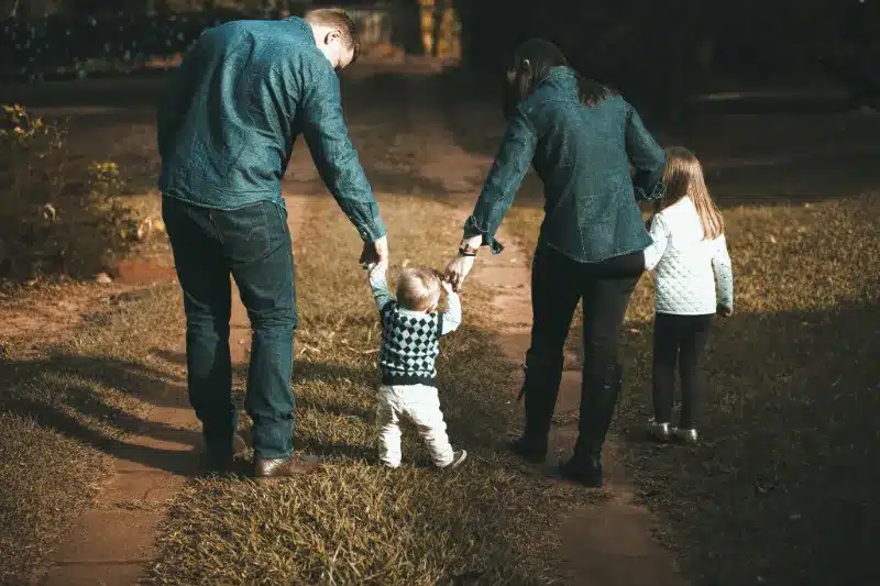 a mother and father holding the hands of a toddler and young girl while walking on a trail outside