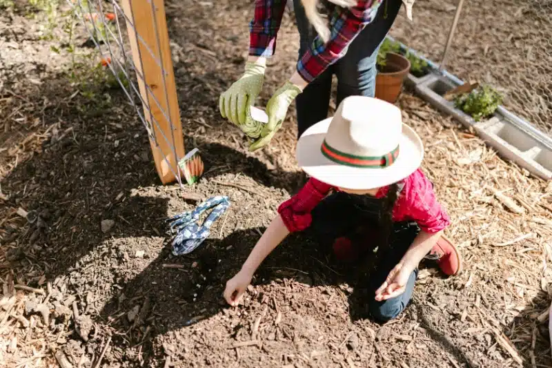 a young boy in a hat and overalls plants a seed in the dirt while his mother helps standing beside him